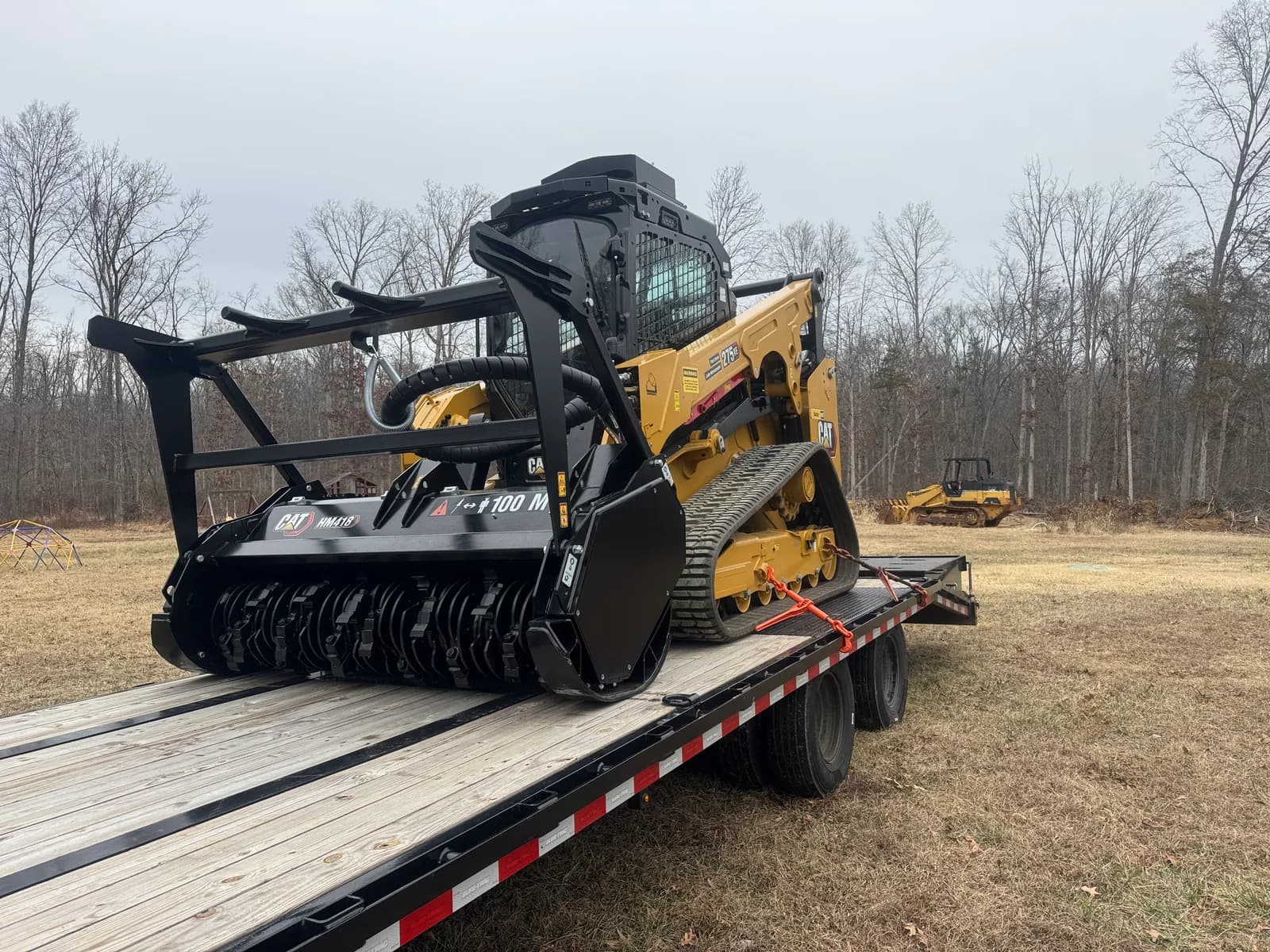 Tracked loader with a forestry mulching head and visible cutting drum on a transport trailer